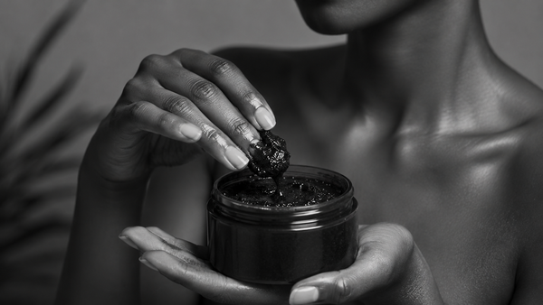 Black and white close-up of a woman holding a jar of African Black Soap paste and gently scooping the natural cleanser with her fingertips.